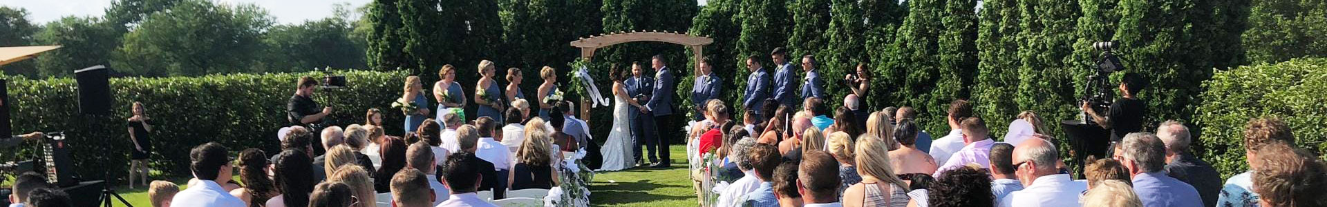 Outdoor wedding ceremony with guests seated, a couple exchanging vows under an arch, surrounded by greenery.