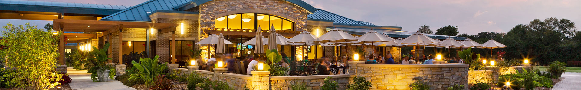 Exterior view of a restaurant with outdoor seating, umbrellas, and lush landscaping during twilight.