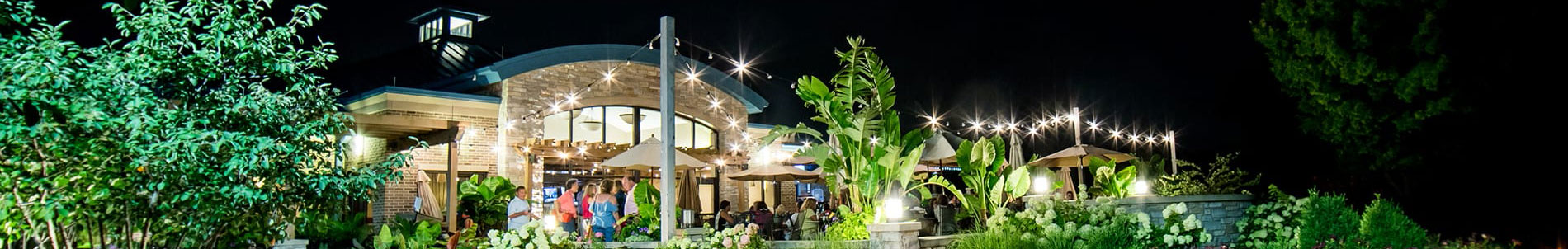 A lively outdoor dining area at night, illuminated by string lights, surrounded by lush greenery and plants.