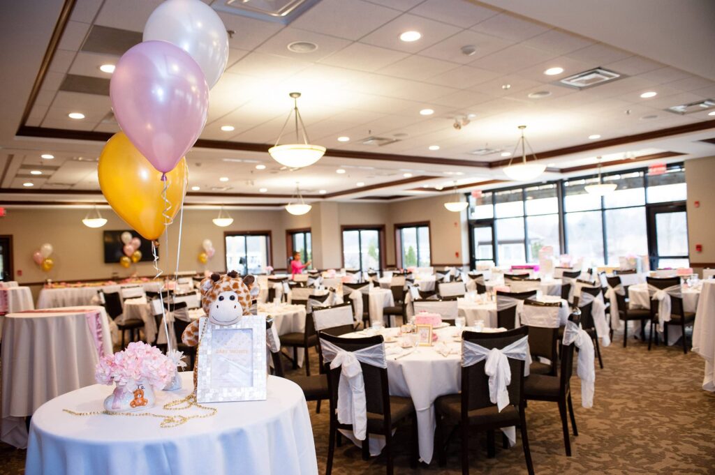 Decorated banquet hall for a baby shower, featuring balloons, a stuffed giraffe, and floral arrangements.