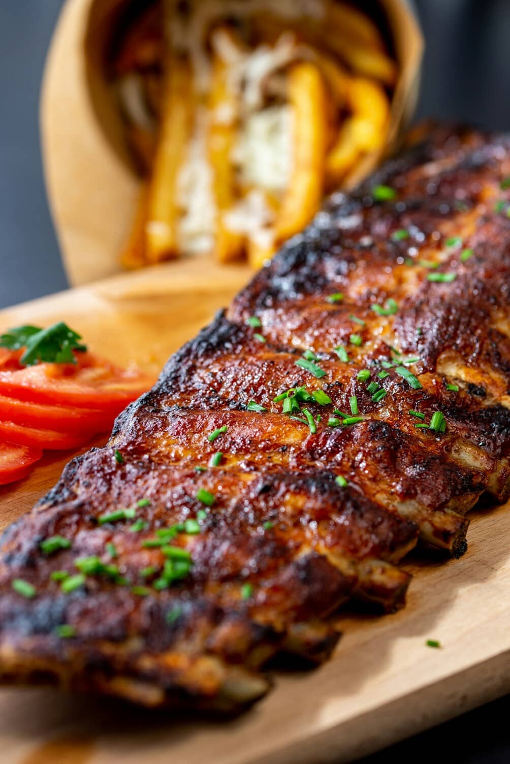 Grilled ribs garnished with chives and served with sliced tomatoes and fries in a paper container.