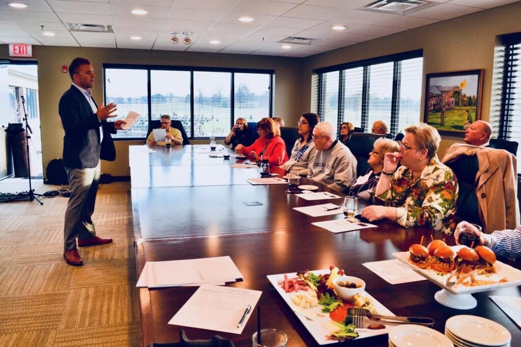 A speaker presents to an attentive audience at a corporate event, with a table of food in the foreground.