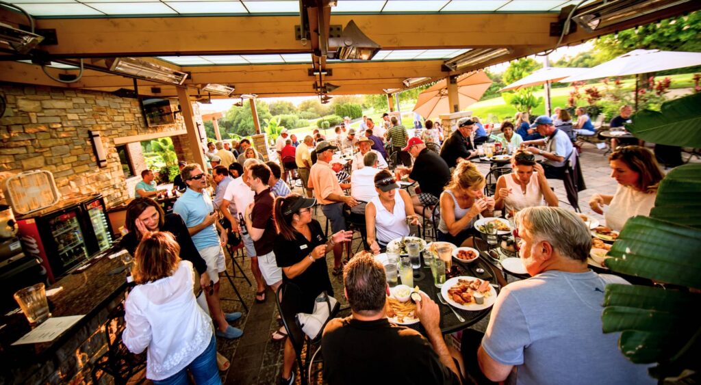 A lively outdoor dining scene with people enjoying meals and drinks at tables under a patio.