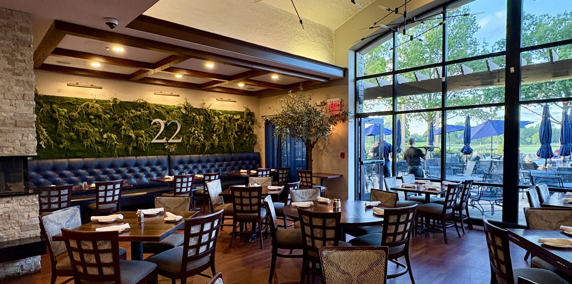 Interior of a restaurant featuring a green moss wall, blue seating, and large windows with outdoor seating visible.